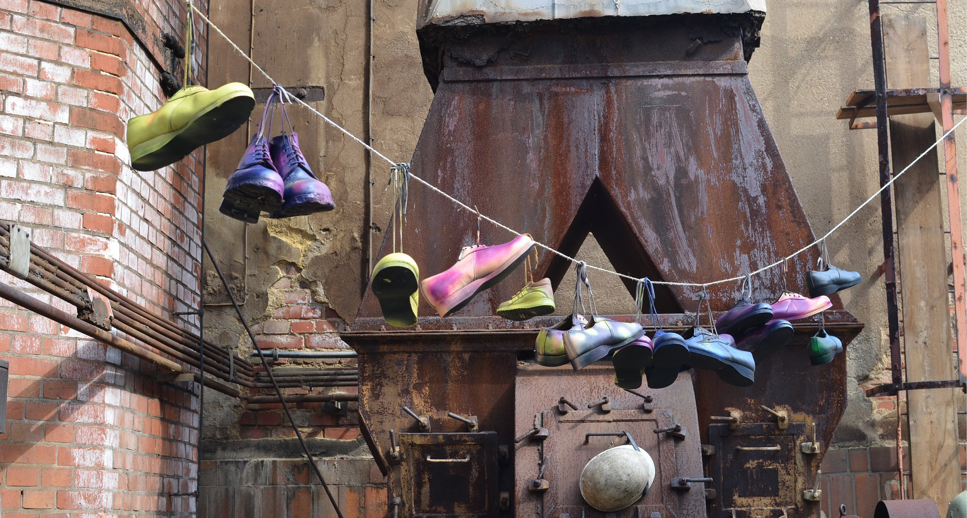 Auf der Abbildung sieht man Teile eines alten Industriestandortes. Vor den rostigen Maschinen sind mehrere bunt lackierte Schuhe nebeneinander an einer Leine aufgehängt.  