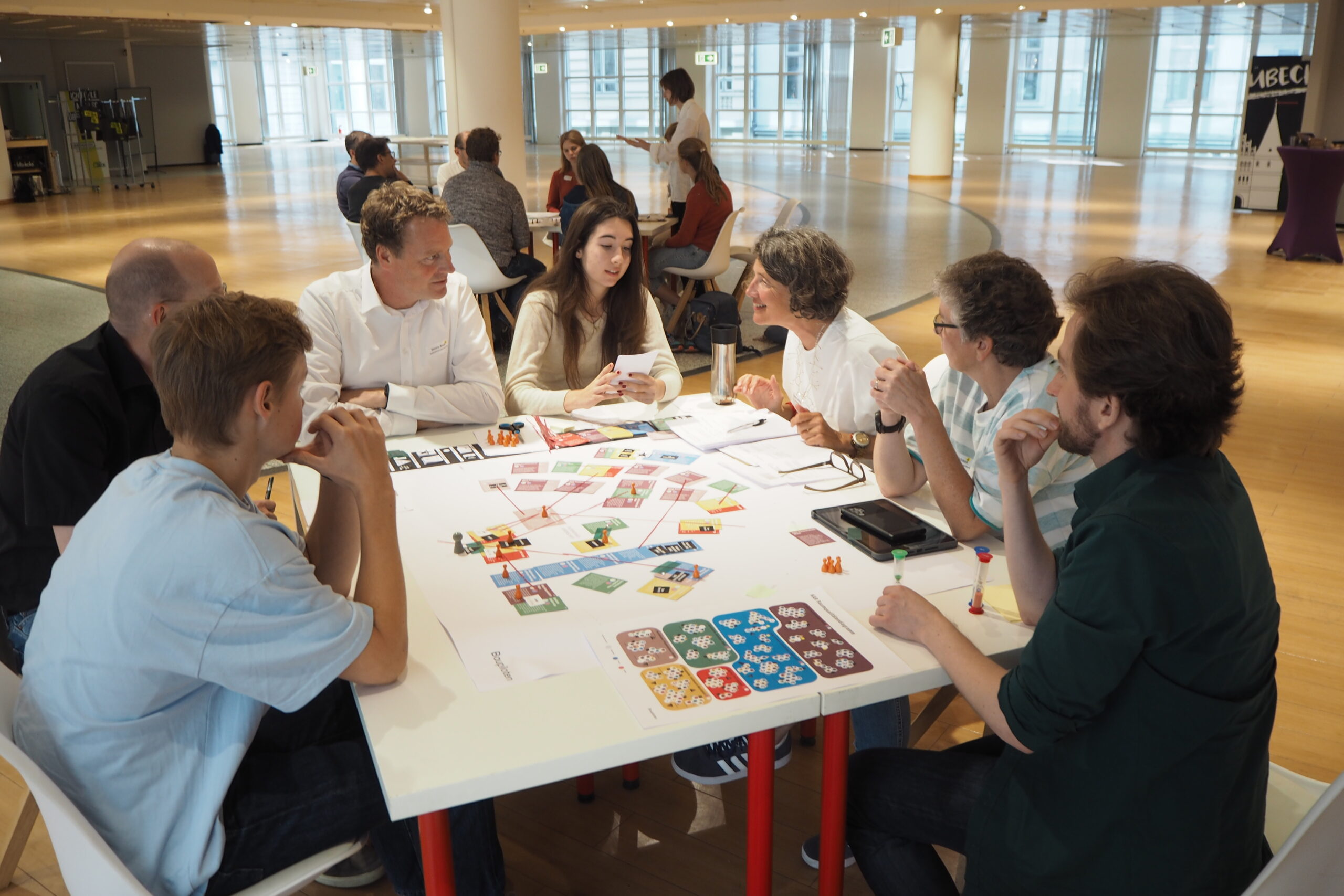 Eine Gruppe von Menschen sitzt an einem Tisch und spielt ein Planspiel. Im Hintergrund eine zweite Gruppe.