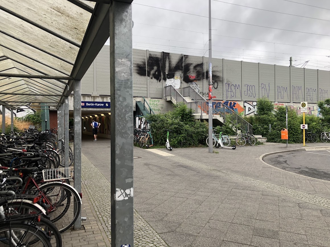 Unterführung zum Bahnhof Berlin-Karow mit Fahrradabstellanlage auf der linken Seite und einer Bushaltestelle auf der rechten Seite. Vor dem Bahnhof sind zahlreiche Fahrräder und vereinzelt E-Scooter abgestellt. Der Blick auf die Gleise wird durch eine Schallschutzwand verdeckt. Eine Treppe führt hinauf zur Schallschutzwand.