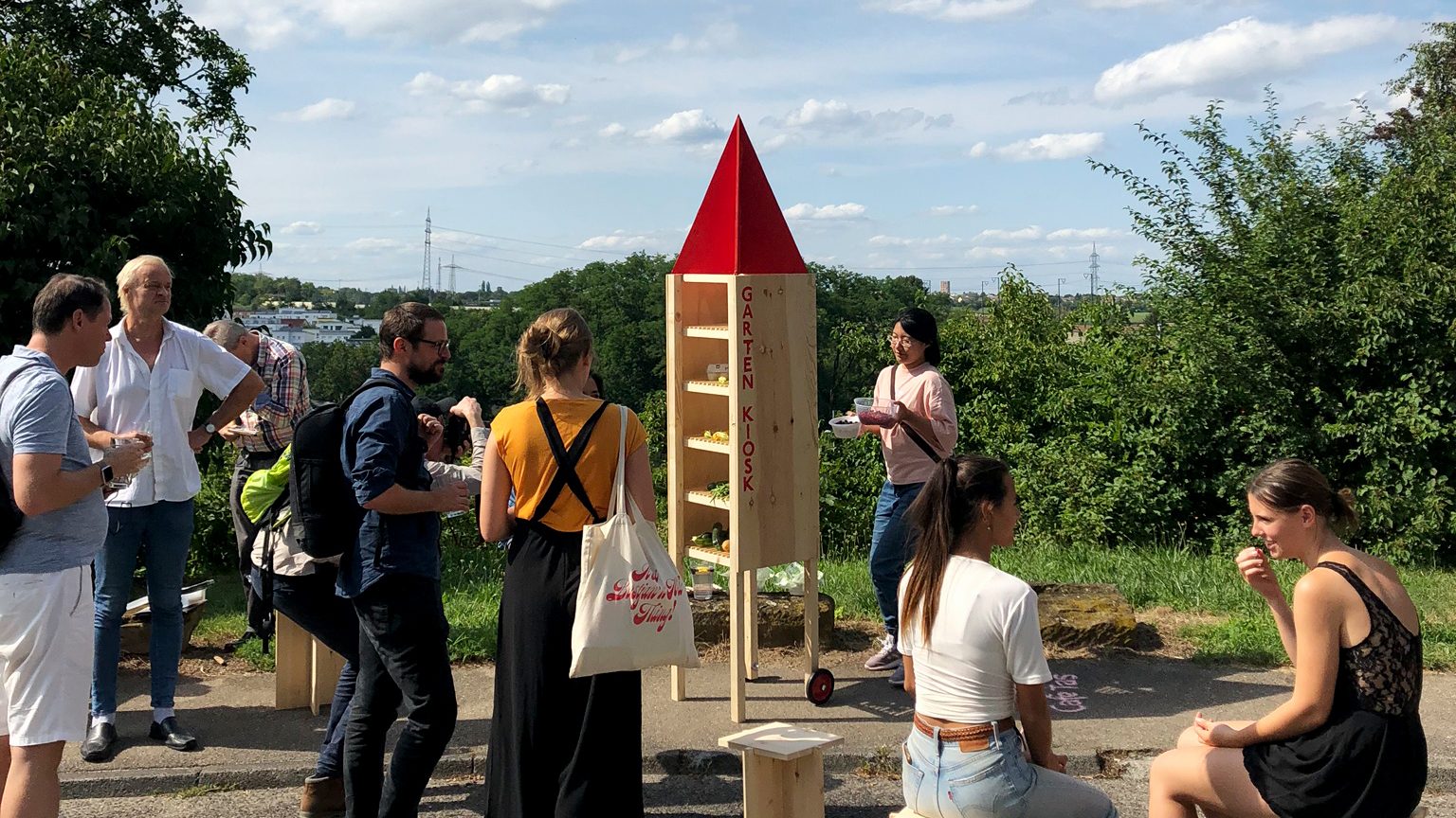 Das Foto zeigt eine Aktion des Labor Böckinger Straße im öffentlichen Raum zur Steigerung der Aufmerksamkeit und des Spaßfaktors der Beteiligung. Es sind Bürger:innen unterschiedlicher Altersgruppen zu sehen, die in freier Natur um ein Holzobjekt mit der Aufschrift „Garten-Kiosk“ stehen und sitzen.