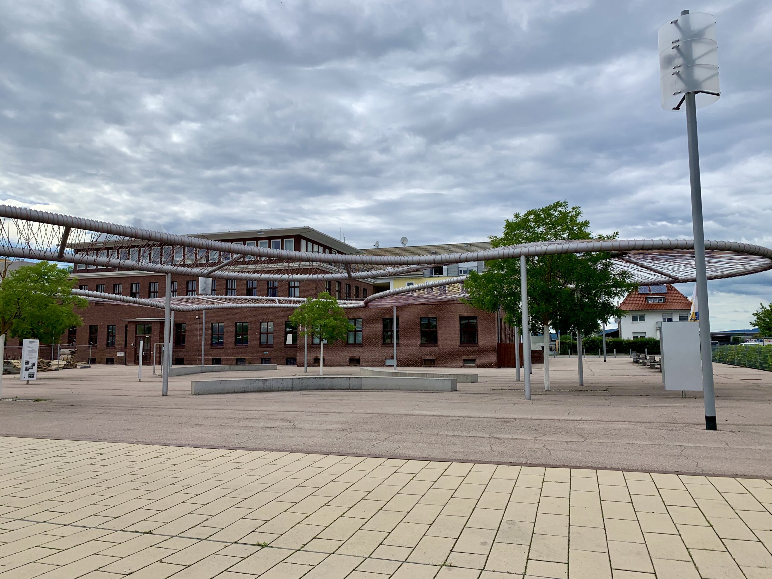 : Neugestalteter Quartiersplatz mit Sitzgelegenheiten, Infotafeln, Bäumen, Leuchten und einer Kunstinstallation, die Bezug auf die ehemalige Kunstseidenproduktion nimmt, im Vordergrund und denkmalgeschützte Backsteinbauten der ehemaligen ENKA-Hauptverwaltung im Hintergrund.