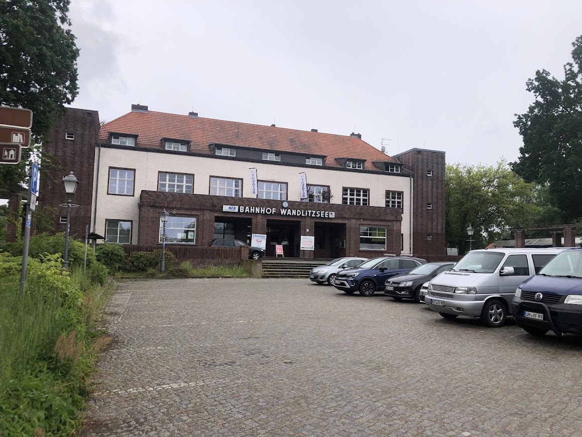 Bahnhofsgebäude mit Treppenaufgang und davorliegendem Parkplatz, der durch PKWs belegt ist.