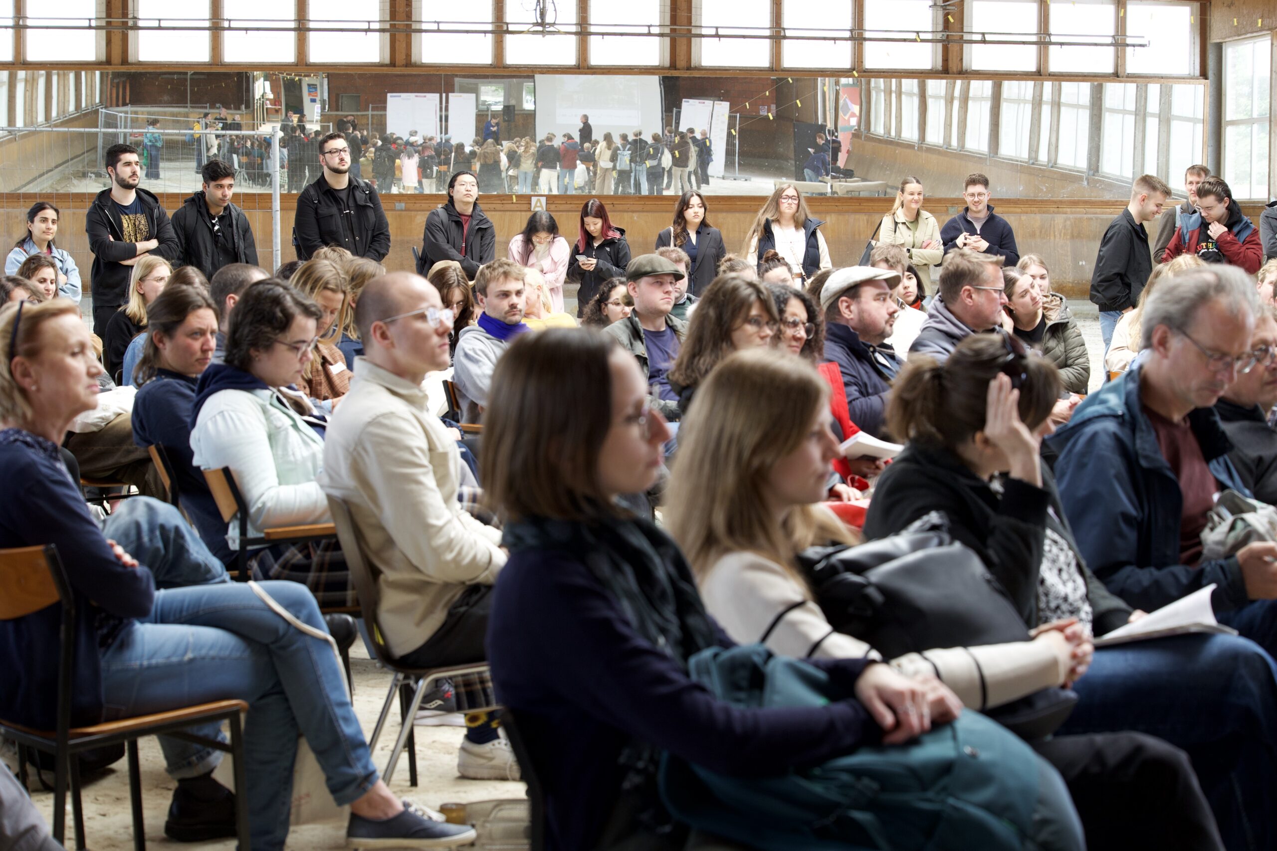 Das Foto zeigt die Zuhörer:innenschaft eines Vortrags. Knapp 20 Personen sitzen auf Stühlen in Reihen. Dahinter stehen circa 10 Personen nebeneinander in einer Reihe. Im Hintergrund reflektiert sich die Szene in einer Spiegelwand.