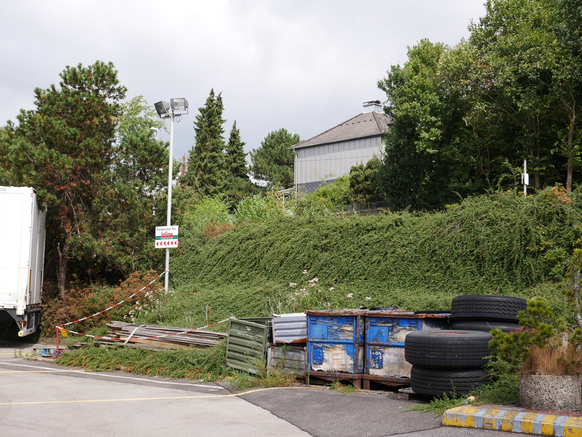 Eine ungenutzte begrünte Hangfläche, davor Baumaterialien und Parkplätze.