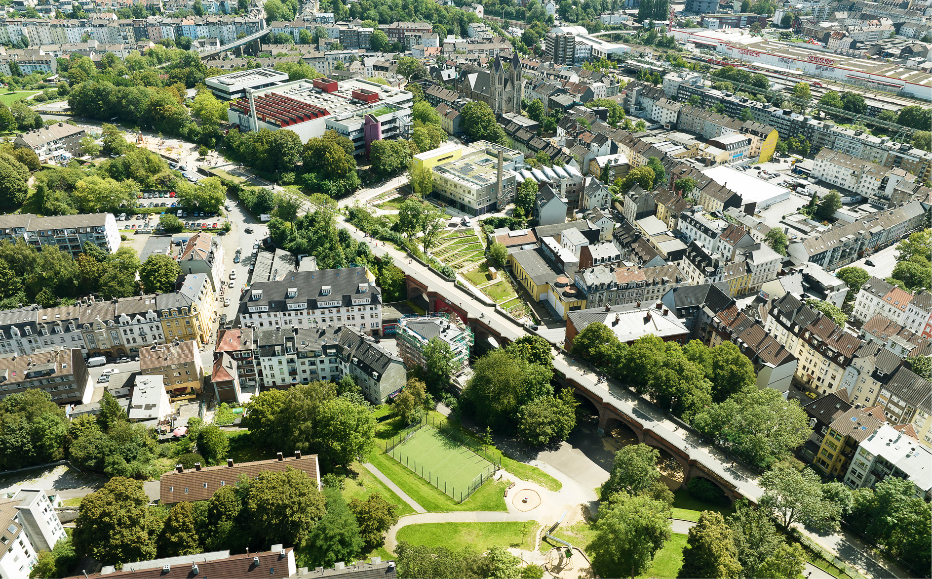 Der Stadtteil Wuppertal-Oberbarmen aus der Luft. Im Zentrum ist der BOB CAMPUS mit dem Nachbarschaftspark zu sehen, daneben das Schulzentrum Ost. Das Hauptgebäude des Campus aus den 1970er Jahren sticht durch die lichtdurchlässige Fassade aus Polycarbonat und den gelben Erschließungsbügel hervor. Foto: Michel Wenzel