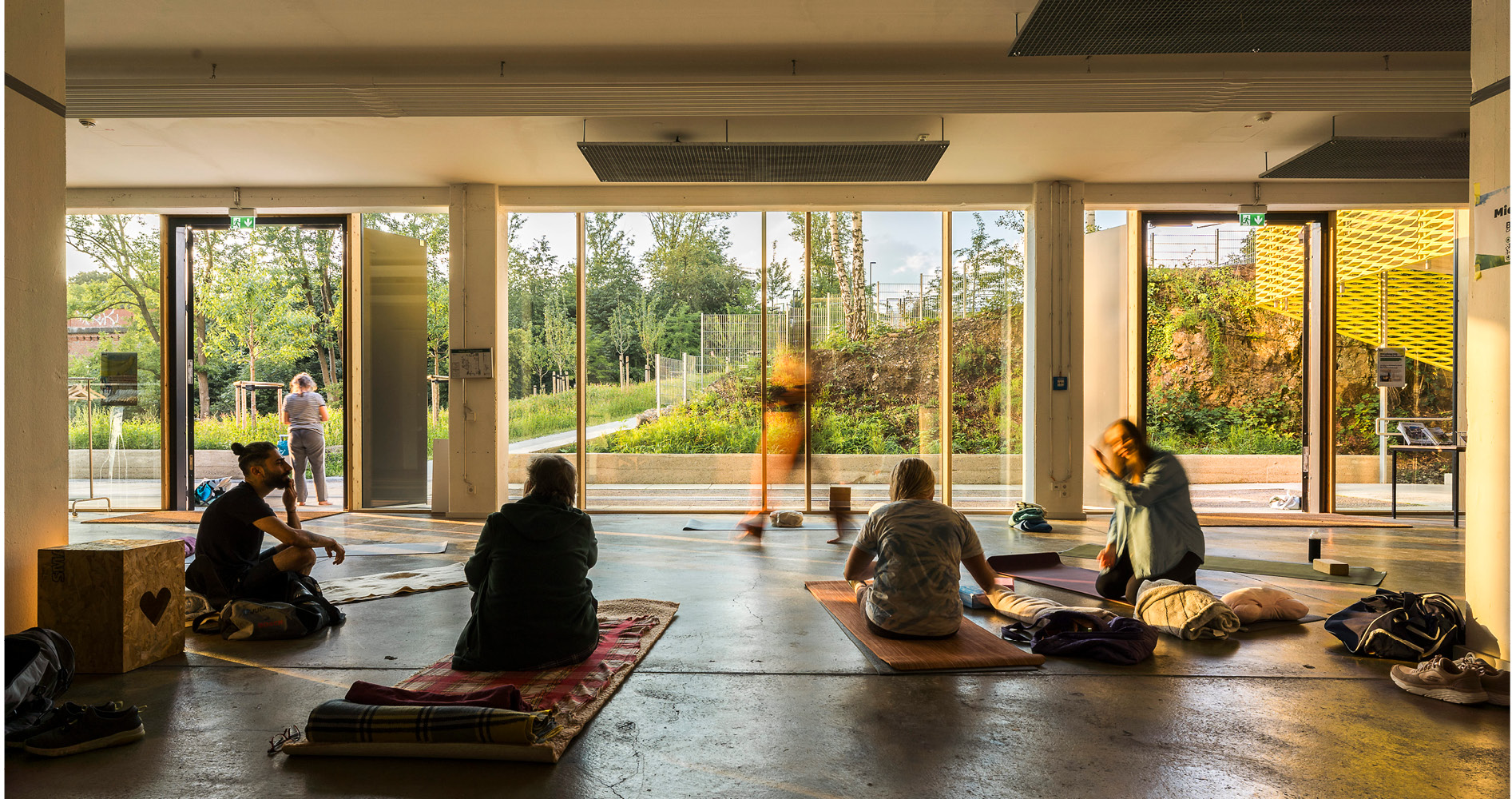 In der Nachbartschaftsetage sitzen Teilnehmer:innen eines Yoga-Kurses auf ihren Yogamatten. Durch die großen Fenster der Nachbarschaftsetage sichtbar befindet sich im Hintergrund der Nachbarschaftspark in der Abendsonne. Foto: Simon Veith